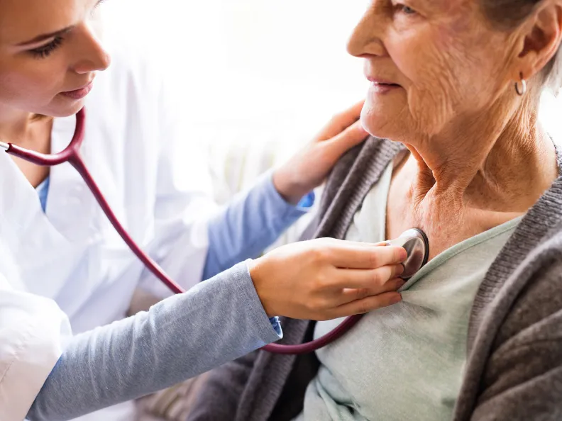 A healthcare professional uses a stethoscope to listen to an elderly person’s chest during a medical examination. The person is wearing a light green top and gray cardigan, and both appear calm.