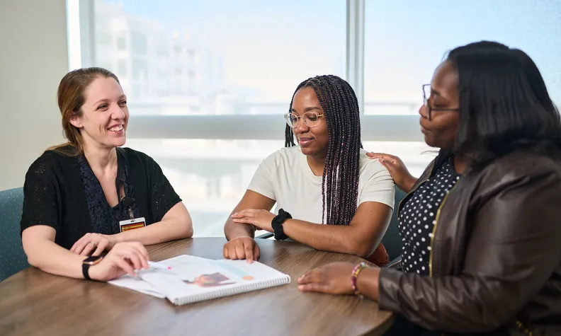 Three people seated at a round table in a bright room, having a discussion, referring to paperwork on the table. One is in medical scrubs, the other two are not.
