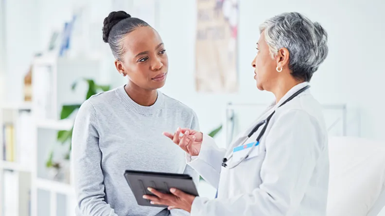 A doctor in a white coat holds a tablet and talks with a smiling patient sitting on an exam table in a bright medical office.