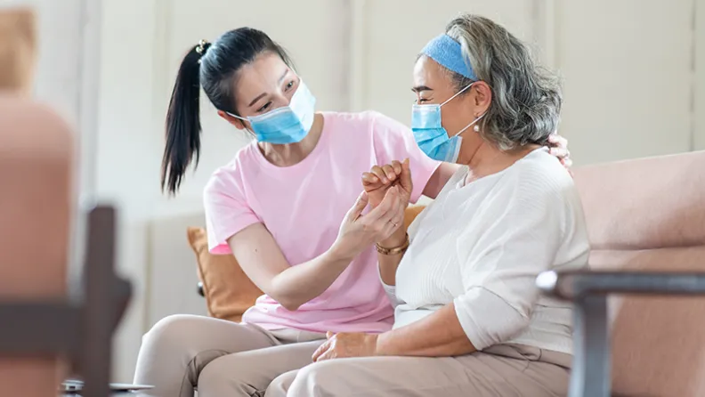 A caregiver gently supports and holds the hand of an older adult seated on a couch.
