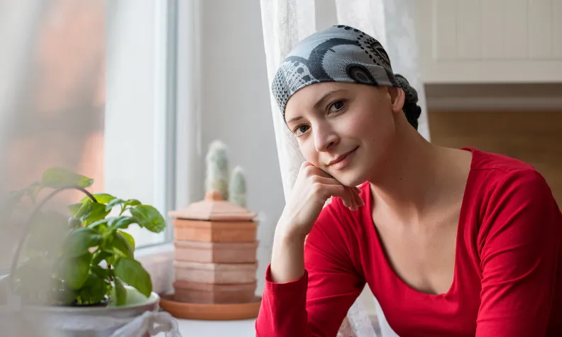 A person wearing a patterned headscarf sits by a bright window, resting their head on one hand. 