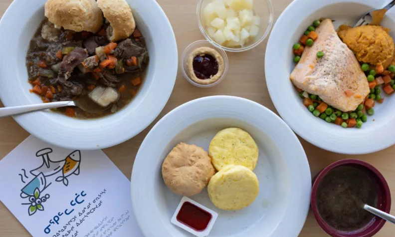 Vue de dessus d’un repas hospitalier comprenant des mets traditionnels, dont un ragoût avec du bannock, du saumon avec des légumes, une soupe et des accompagnements disposés sur une table.