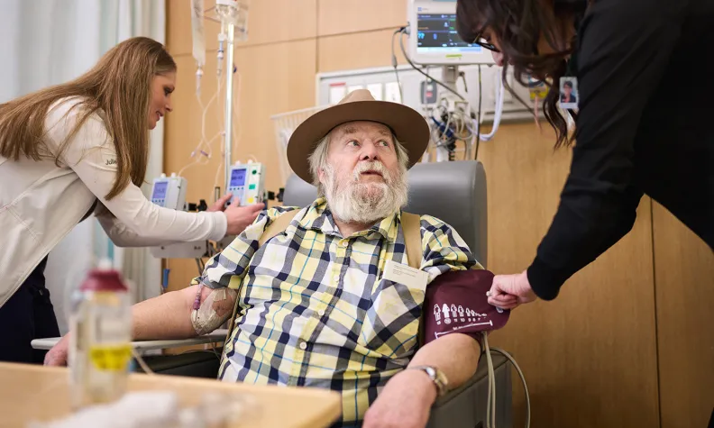 An older person in a plaid shirt and hat sits in a medical chair while two healthcare workers take blood pressure and adjust an IV.