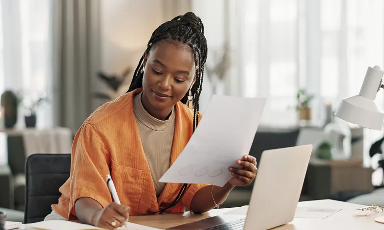 Person sitting at a desk reviewing a document while working on a laptop in a bright office setting.