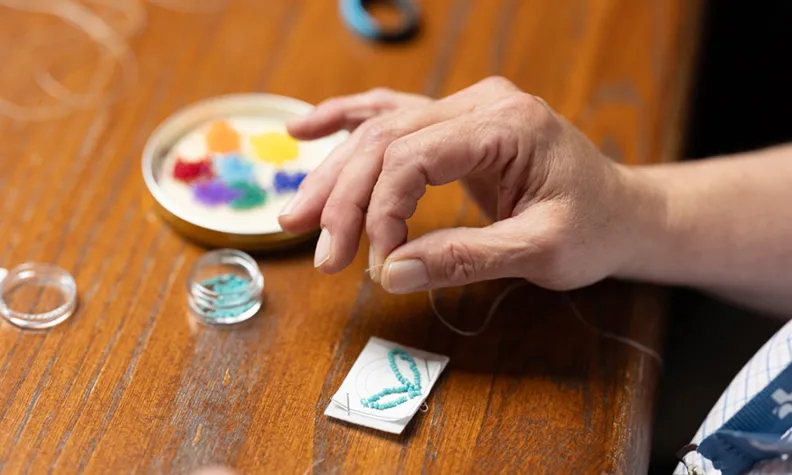 A close-up shot of a workshop participant’s hand, fingers gripping the end of a strand of white thread. Piles of small, colourful beads can be seen on the table, as well as the early stages of a beaded keychain. 
