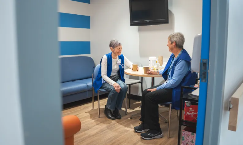 Through a doorway, a view of volunteers Barb McDonald and Moninna Running sitting at a table and laughing.