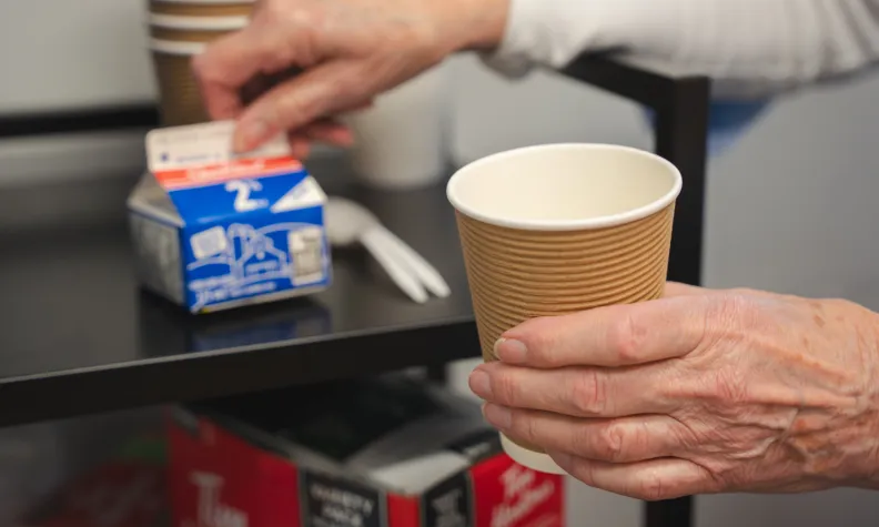 A close-up of an older woman's left hand holding a cup of coffee. Behind it, her right hand holds a container of milk.
