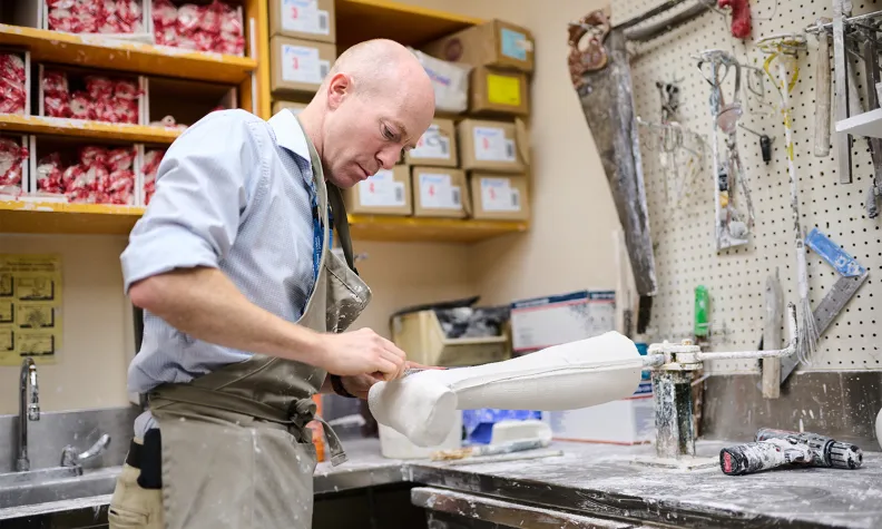 A person in an apron works on a white plaster leg cast in a workshop, surrounded by tools, shelves with boxes, and plaster dust on the counter.