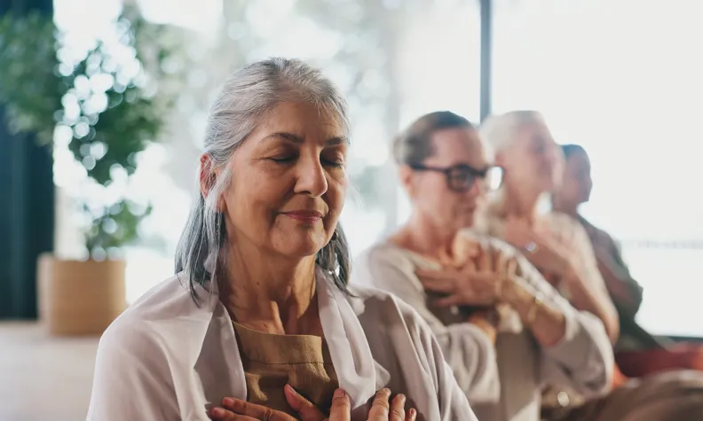 Person seated with eyes closed and hands on chest, participating in a calm group wellness or meditation session.