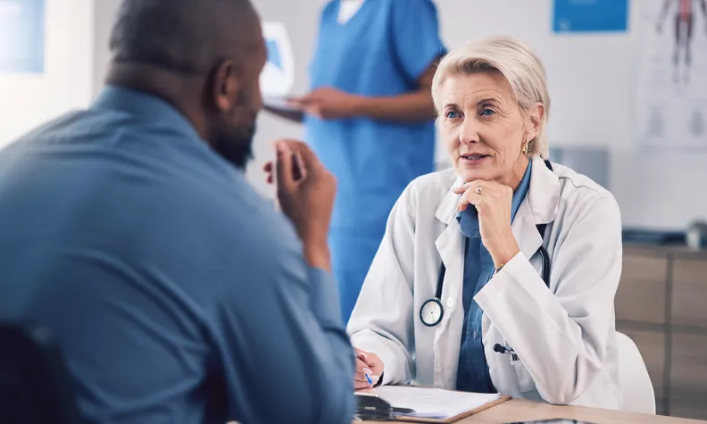 Close-up of a healthcare professional seated at a desk, listening and speaking with a patient during a consultation.
