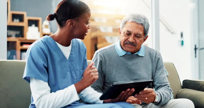 Healthcare worker sits with an older adult, reviewing information together on a tablet in a home setting.