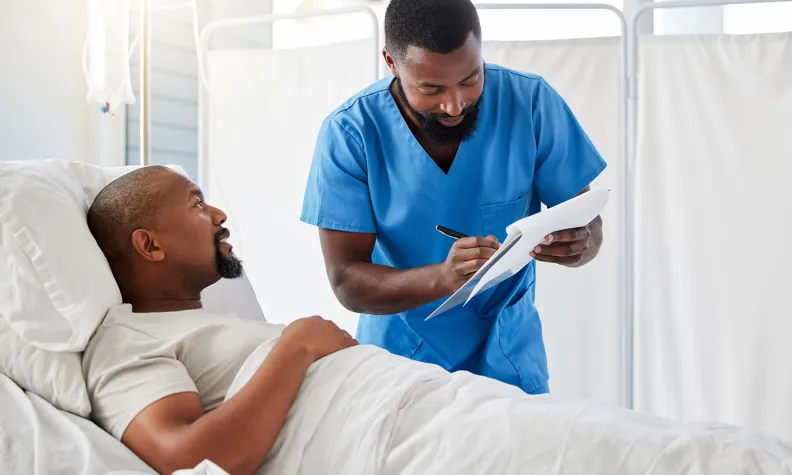 Patient lying in a hospital bed while a healthcare worker in scrubs writes notes on a clipboard beside them.