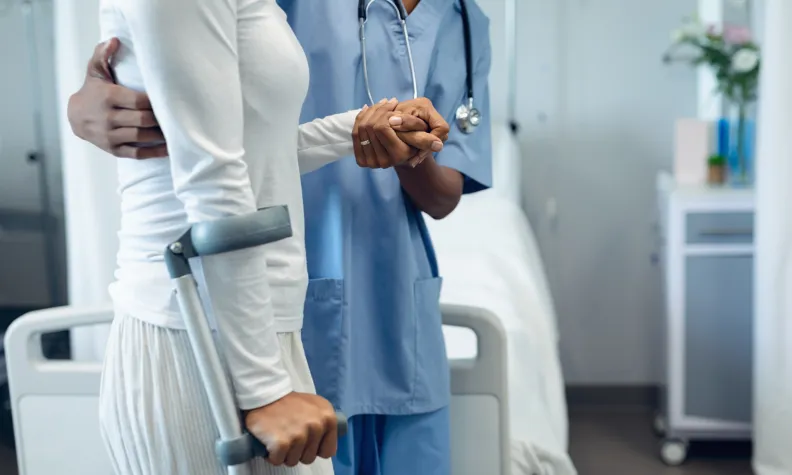 Healthcare provider assists a patient using crutches beside a hospital bed during discharge preparation.