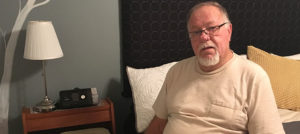 Ron Cooper is sitting on his home bed