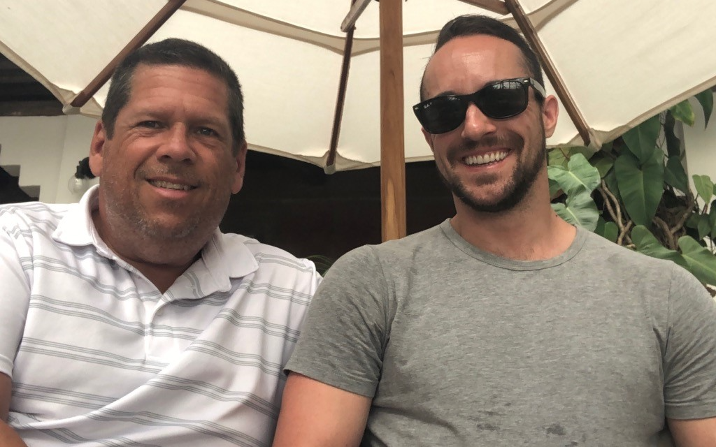 Stuart MacMillan (left) and his husband, Sébastian LeBel (right), sitting under a white umbrella