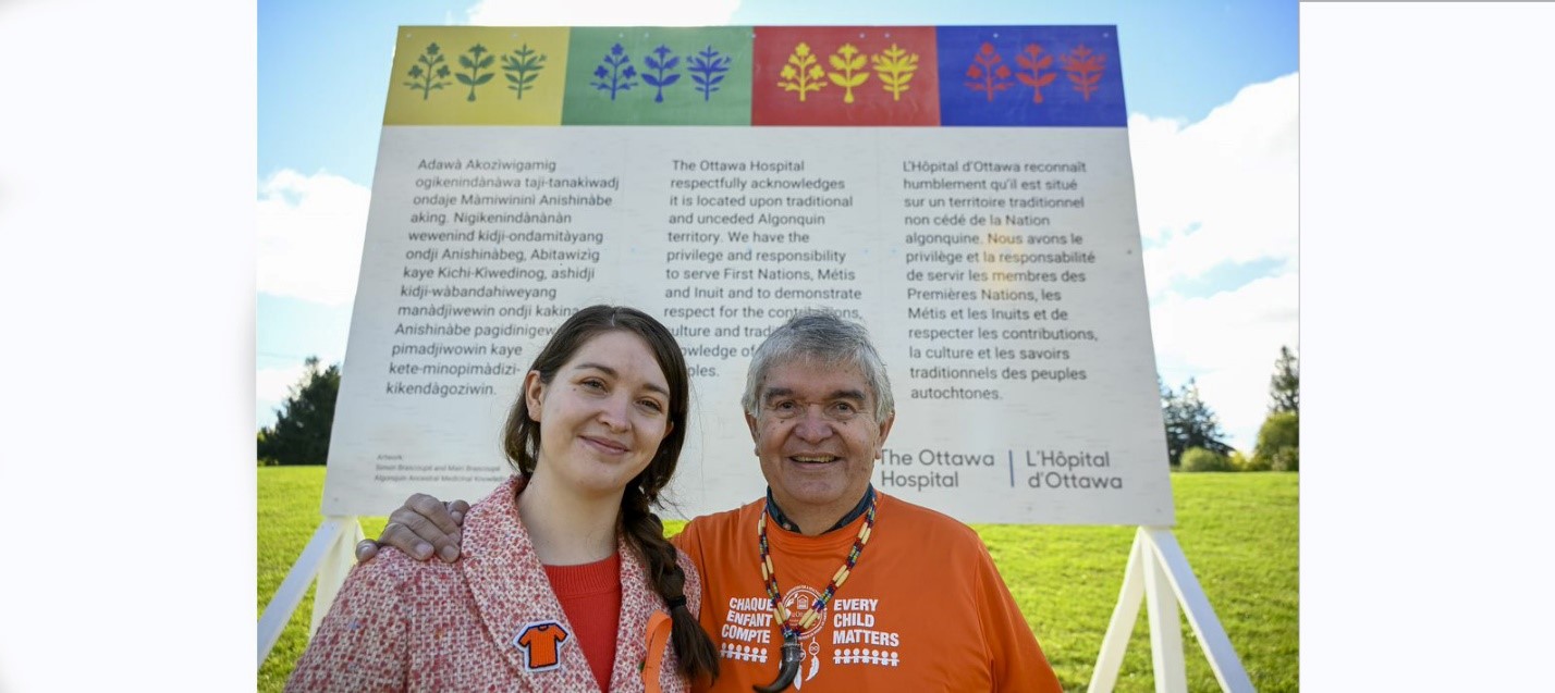 Mairi Brascoupé (left) and Simon Brascoupé standing in front of the land acknowledgement sign that features their original artwork.
