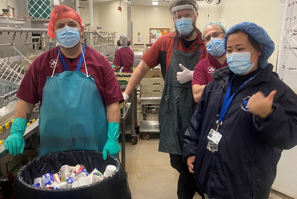 Four staff members stand in front of large containers full of recyclable material.