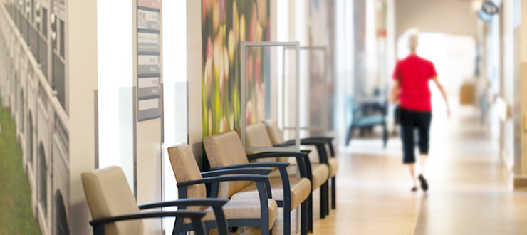 A wide view of a hallway at The Ottawa Hospital. Chairs are lined up against a wall on the left of the hallway, and in the distance, a person is walking towards an exit.