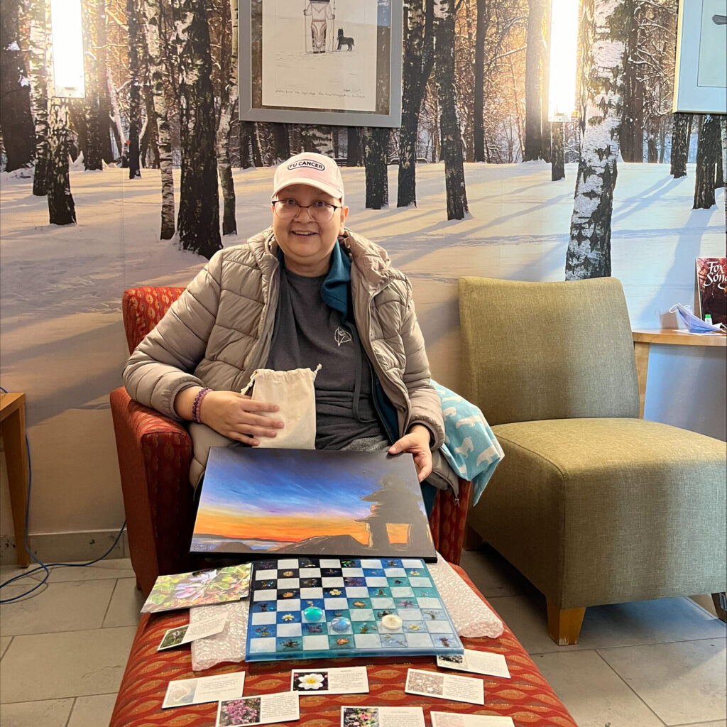 Photo de Jennifer Burmingham, patiente atteinte d’un cancer, assise sur une chaise, souriant et tenant dans ses mains une peinture et un petit sac, avec un ensemble de damier en résine coulée et de petites cartes posés sur un pouf devant elle.  