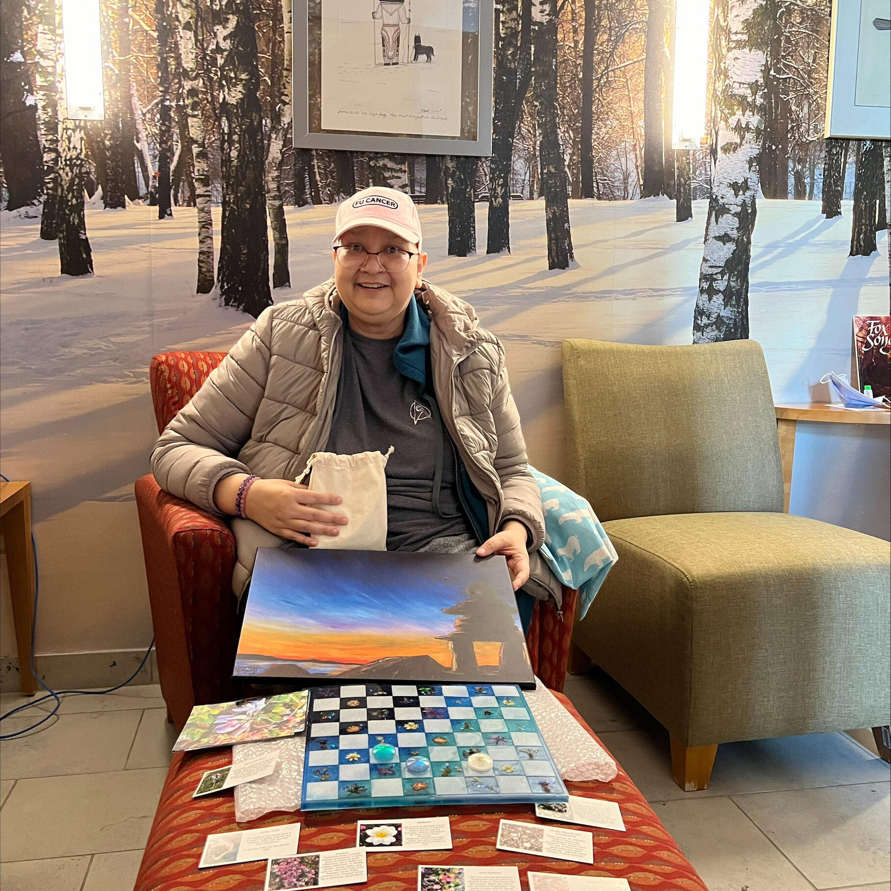 Photo of Jennifer Burmingham, a cancer patient, smiling and sitting in a chair while holding a painting and small bag, with a resin-cast checkerboard set and small cards on an ottoman in front of her.