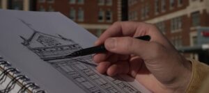 Close-up of an artist’s hand holding a sketch book, sketching the Civic Campus with a black market.