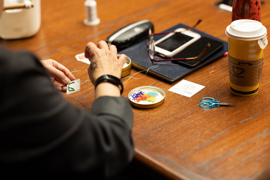 Une participante à l’atelier cousant des perles vertes sur son patron. Sur l’espace de travail devant elle, des perles, des ciseaux, du fil blanc, un bloc-notes, des lunettes, des téléphones et une tasse de thé réutilisable.