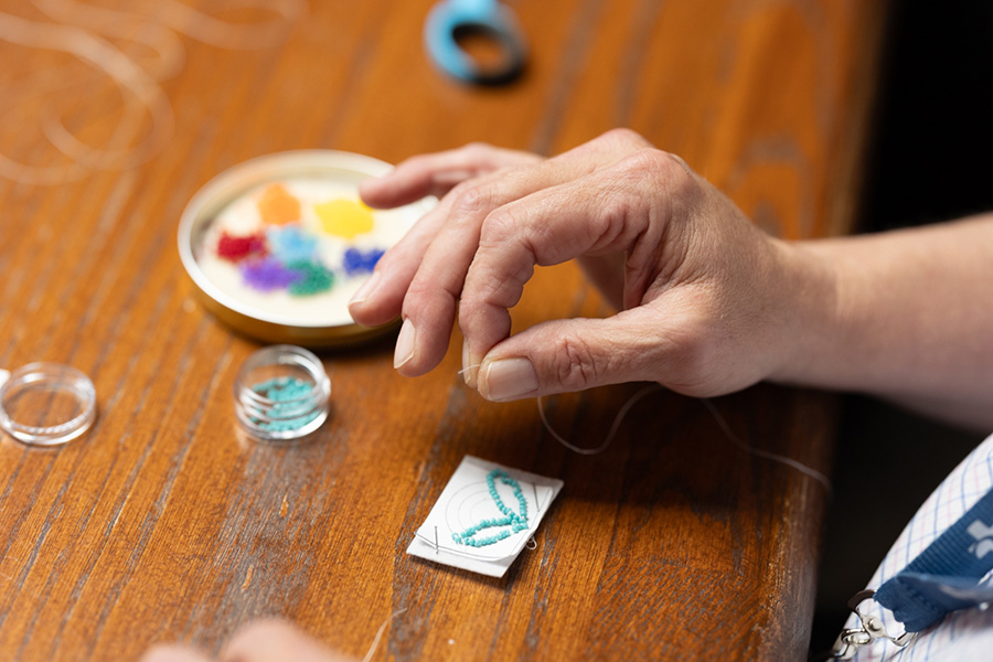 Gros plan de la main et des doigts d’une participante à l’atelier attrapant un bout de fil blanc. Des petites perles de différentes couleurs s’amoncellent sur la table. On y découvre les premières étapes de ce qui deviendra une chaîne porte-clés en perles. 