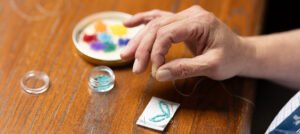A close-up shot of a workshop participant’s hand, fingers gripping the end of a strand of white thread. Piles of small, colourful beads can be seen on the table, as well as the early stages of a beaded keychain.