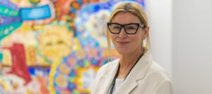 Dr. Mary Marquardt stands in front of some colourful, therapeutic artwork created by clients in the OnTrack program.