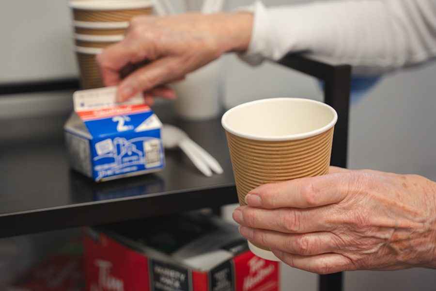A close-up of an older woman's left hand holding a cup of coffee. Behind it, her right hand holds a container of milk.